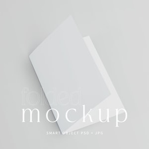 以下が含まれることがあります： 白く折りたたまれたモックアップで、わずかに開いています。背景は明るいグレーです。「folded」という単語は柔らかくエレガントなフォントで表示され、その下に「mockup」があります。「SMART OBJECT PSD + JPG」というテキストが下にあります。