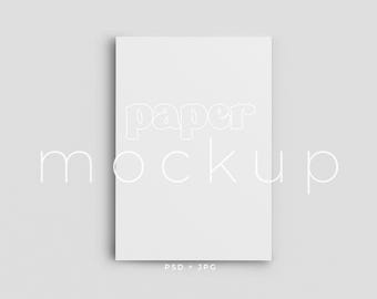 May include: A white paper mockup with the words "paper mockup" in a stylized font. The image is a flat lay on a light gray background. The bottom of the image has the text "PSD + JPG".