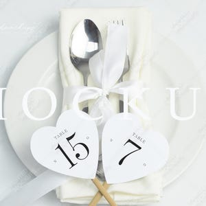 May include: White heart-shaped table number cards with black text reading "Table 15" and "Table 7" on a white background. The cards are placed on a white napkin with a silver fork and spoon tied with a white ribbon.