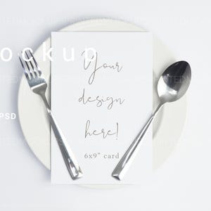 Puede incluir: Una tarjeta blanca de 6x9" con el texto "Your design here!" está centrada en un plato blanco, flanqueada por un tenedor y una cuchara. El tenedor y la cuchara son plateados. La imagen es una maqueta.