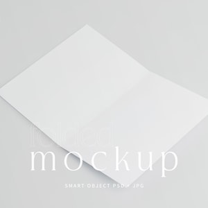 Può includere: Un mockup piegato bianco su sfondo grigio chiaro. Il testo "folded mockup" è visualizzato in un carattere delicato. La parte inferiore dell'immagine include il testo "SMART OBJECT PSD + JPG".