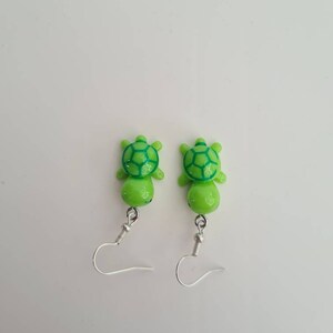 Puede incluir: Un par de pendientes de tortuga verde con ganchos plateados. Las tortugas tienen un caparazón verde con un patrón verde más oscuro.