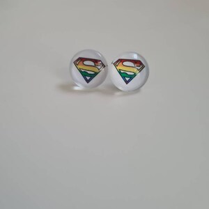 Puede incluir: Un par de pendientes de botón con el logotipo de Superman arcoíris. Los pendientes están hechos de vidrio transparente con un logotipo de Superman arcoíris impreso en la parte delantera.