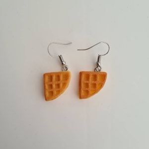 May include: A pair of silver dangle earrings with a waffle-shaped charm. The charms are a light brown color and have a grid pattern.