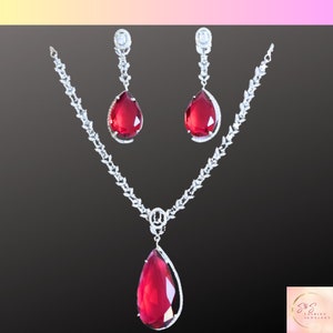 May include: A silver necklace and earrings set with a large red teardrop-shaped gemstone. The necklace features a delicate chain with small clear stones and a pendant with a larger red gemstone. The earrings are dangle earrings with a similar design.