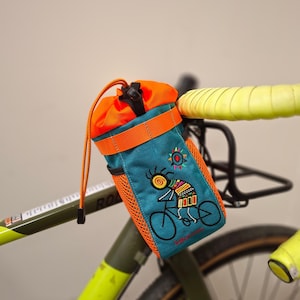 May include: Teal and orange bicycle handlebar bag with an embroidered design of a person riding a bicycle. The bag has a drawstring closure and is attached to the handlebars of a bicycle with yellow tape.