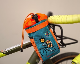 Pode incluir: Bolsa de guiador de bicicleta turquesa e laranja com um desenho bordado de uma pessoa a andar de bicicleta. A bolsa tem um fecho de cordão e está presa ao guiador de uma bicicleta com fita adesiva amarela.