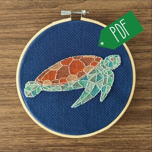 Op de afbeelding: Een geometrisch kruissteekpatroon van een zeeschildpad op een blauwe stoffen achtergrond in een houten borduurring. De tekst PDF is zichtbaar op een groen label.