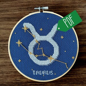 May include: A blue and white embroidery hoop with a gold cross-stitch design of the Taurus constellation. The constellation is outlined in white and the stars are gold. The word "Taurus" is stitched in gold below the constellation.
