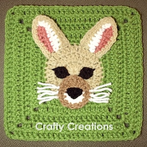 May include: A green crochet square with a brown and white bunny face in the center. The bunny has pink ears, black eyes, a brown nose, and white whiskers. The square has the text "Crafty Creations" at the bottom.
