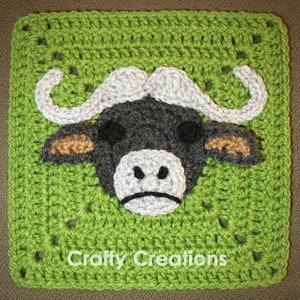 Op de afbeelding: Een groen gehaakt vierkant met een bruin, grijs en wit gehaakt buffelhoofd in het midden. Het vierkant heeft de tekst "Crafty Creations" onderaan.