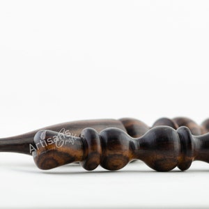 May include: Close-up of a dark brown wooden crochet hook with a pointed tip and a textured handle. The hook is made of a dark wood with a natural grain.