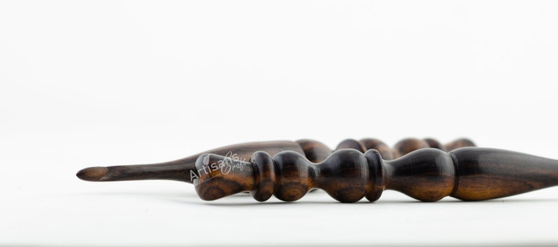 May include: Close-up of a dark brown wooden crochet hook with a pointed tip and a textured handle. The hook is made of a dark wood with a natural grain.