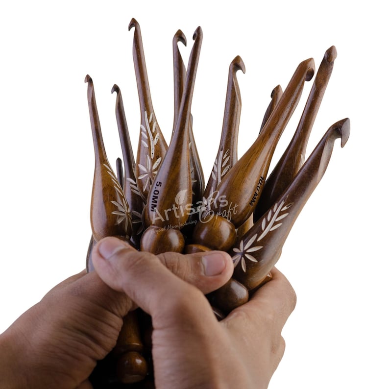 Pode incluir: Um conjunto de agulhas de croch&ecirc; de madeira com desenhos florais entalhados. As agulhas s&atilde;o marrons e t&ecirc;m letras brancas que dizem "5.0MM" e "WWW.OMN.COM".