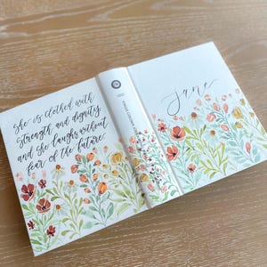 May include: A hardback book with a floral design and the name "Jane" in cursive. The cover features a watercolour illustration of flowers in shades of red, orange, and yellow. The book's spine reads "SINGLE COLUMN JOURNALING". The front cover has the quote "She is clothed with strength and dignity and she laughs without fear of the future."