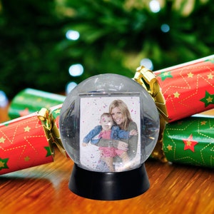 Puede incluir: Un globo de nieve de vidrio transparente con una foto de una mujer y un niño en su interior. El globo de nieve está sentado en una mesa de madera con dos petardos navideños rojos y verdes a cada lado.