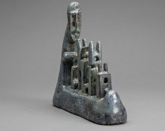 Op de afbeelding: Een blauwgrijze keramische sculptuur met een lange, gestileerde figuur die achter een rij kleinere, abstracte, torenachtige structuren staat. Het object heeft een textuurrijke, gespikkelde glazuurafwerking met donkere vlekjes.