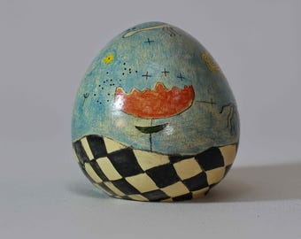 May include: A ceramic egg-shaped sculpture with a whimsical design. The top half is light blue with painted stars, a flower, and other abstract shapes. The bottom half features a black and white checkered pattern.