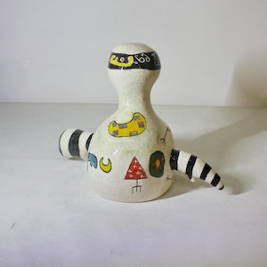 May include: White ceramic sculpture with black and white stripes, yellow, red, blue, and green geometric designs. The sculpture has a rounded head and a long, thin body.