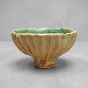 May include: A small, hand-formed ceramic bowl with a textured, fluted exterior in a matte beige finish with subtle reddish-brown highlights. The interior features a vibrant, speckled turquoise glaze that contrasts with the earthy outer surface.
