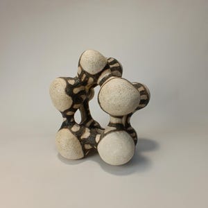 May include: Abstract ceramic sculpture with a cream-colored base and dark brown, organic patterns. The sculpture has a unique, interconnected form with rounded shapes, creating a visually interesting piece of art.