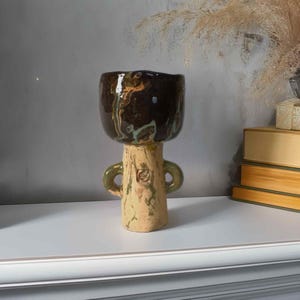 May include: A handmade ceramic vase with a dark brown, textured top and a light tan base. The vase has two small green handles and a unique, organic shape. The vase is a decorative item.