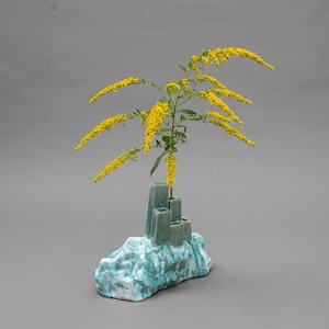 May include: A ceramic vase with a textured, light blue and white base resembling a rock formation, featuring four vertical, rectangular openings of varying heights. A single stem of bright yellow goldenrod flowers is placed in one opening.
