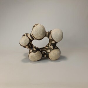 May include: A ceramic sculpture with a unique, organic form. The piece features interconnected, rounded shapes in off-white with a dark brown, patterned design. The sculpture has a textured surface and a central opening, creating an abstract, artistic statement.