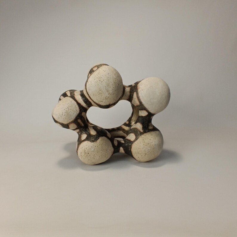 May include: A ceramic sculpture with a unique, organic form. The piece features interconnected, rounded shapes in off-white with a dark brown, patterned design. The sculpture has a textured surface and a central opening, creating an abstract, artistic statement.