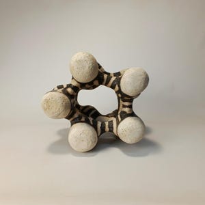 May include: A unique, abstract ceramic sculpture. The piece features a central circular opening, with six rounded, off-white spheres connected by a black and white striped pattern. The sculpture is a decorative art piece.