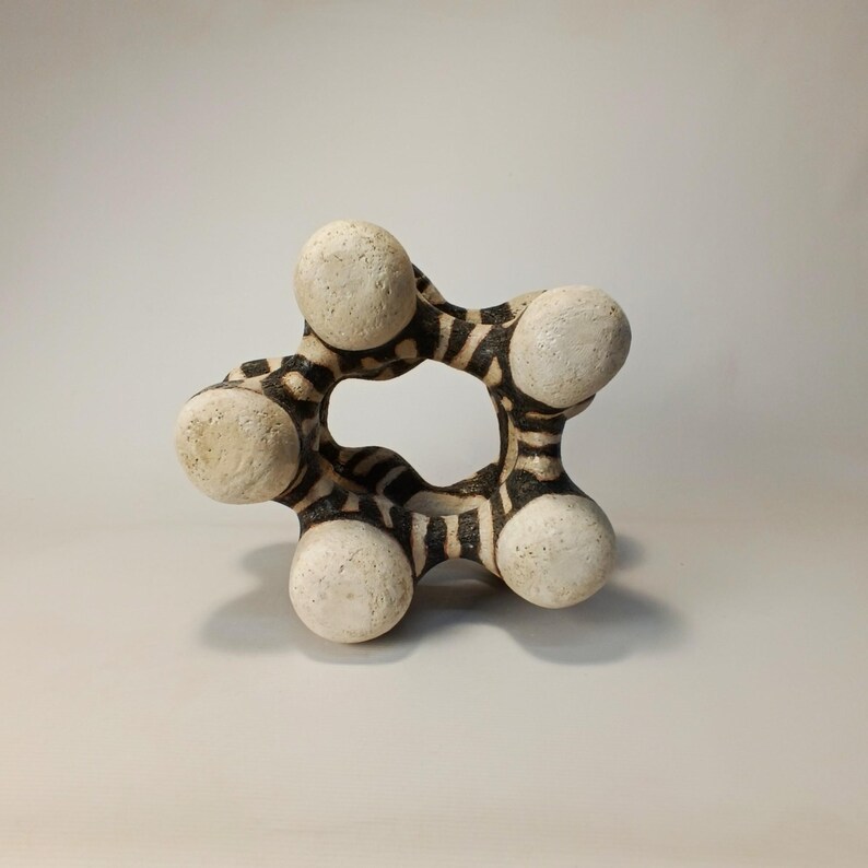 May include: A unique, abstract ceramic sculpture. The piece features a central circular opening, with six rounded, off-white spheres connected by a black and white striped pattern. The sculpture is a decorative art piece.