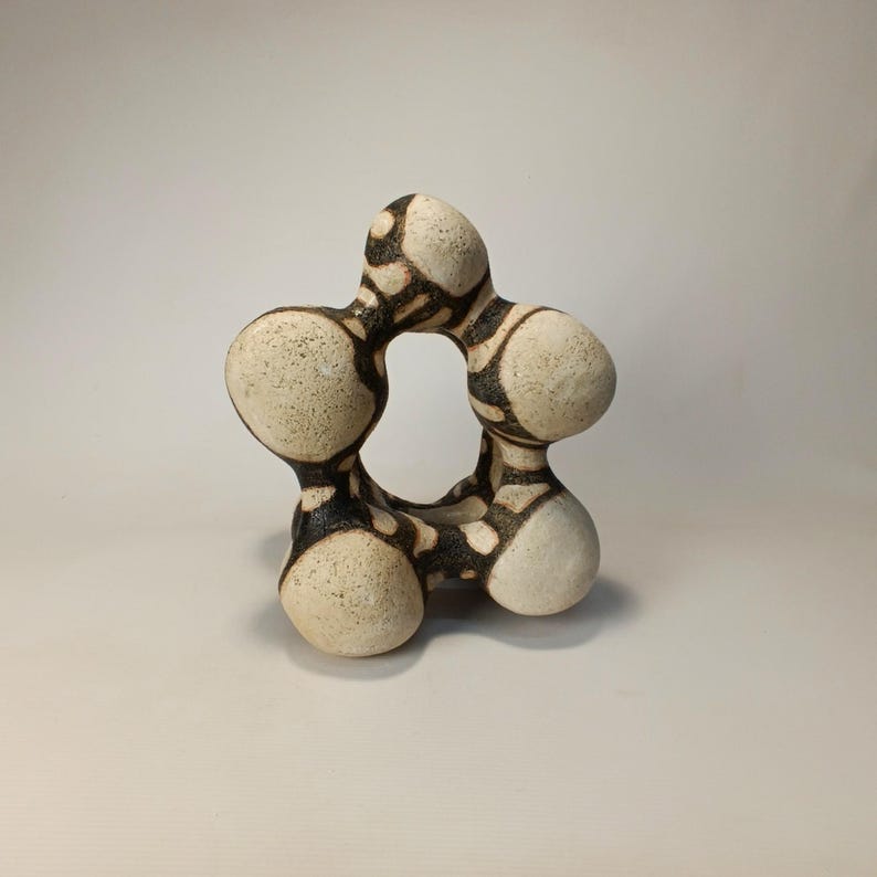 May include: A unique, abstract ceramic sculpture with a star-like shape. The off-white piece features black, organic patterns and a central opening. The textured surface and rounded forms create a modern, artistic aesthetic.