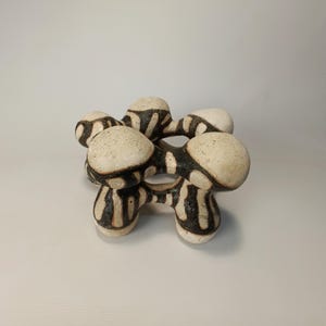 May include: A ceramic sculpture with a unique, abstract design. The piece features interconnected, rounded forms in off-white with black, vertical stripe patterns. The sculpture has a textured surface and is displayed against a plain background.