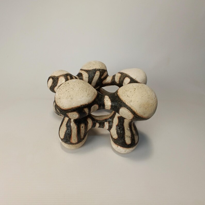 May include: A ceramic sculpture with a unique, abstract design. The piece features interconnected, rounded forms in off-white with black, vertical stripe patterns. The sculpture has a textured surface and is displayed against a plain background.
