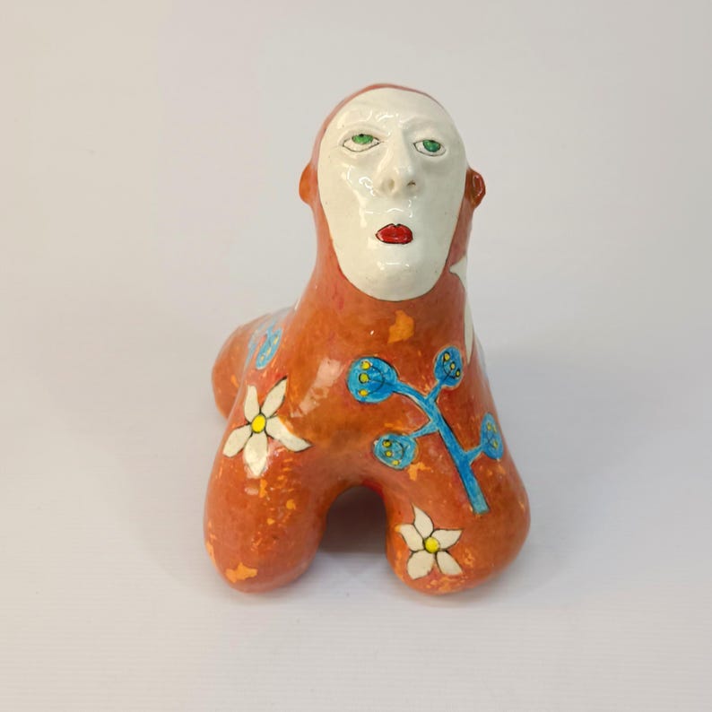 May include: A ceramic sculpture of a stylized human figure with a white face, red lips, and green eyes. The figure is painted in orange, blue, and white with floral designs.
