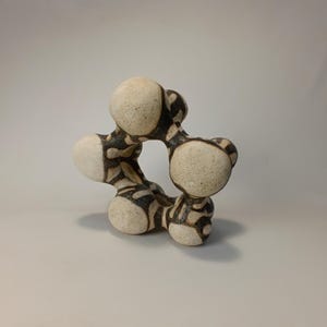 May include: A ceramic sculpture with a unique, abstract design. The piece features a series of interconnected, rounded forms in off-white with a black, patterned overlay. The sculpture has a central opening and a textured surface, suitable for home decor.