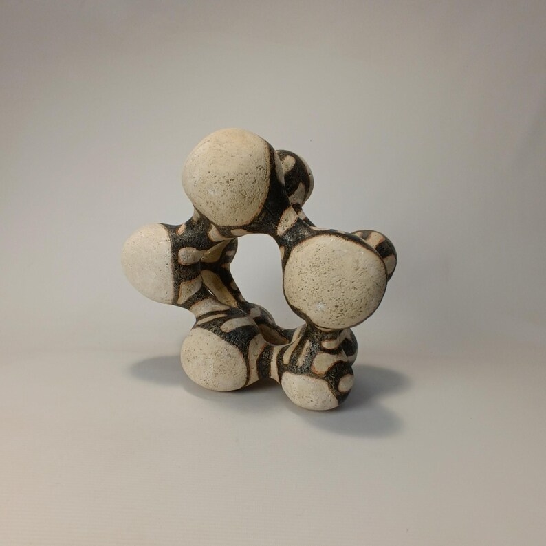 May include: A ceramic sculpture with a unique, abstract design. The piece features a series of interconnected, rounded forms in off-white with a black, patterned overlay. The sculpture has a central opening and a textured surface, suitable for home decor.