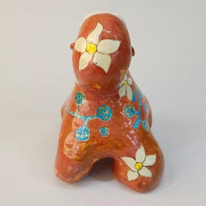 May include: A ceramic sculpture of a stylized figure with a red, orange, and yellow glaze. The figure is decorated with white flowers and blue and yellow vines.