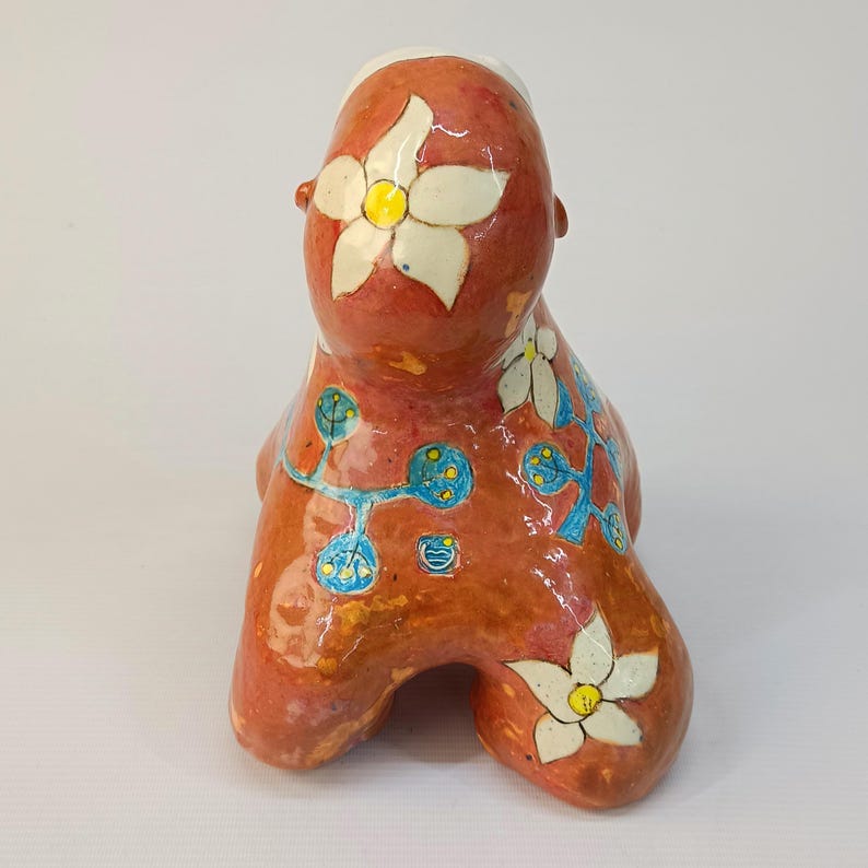 May include: A ceramic sculpture of a stylized figure with a red, orange, and yellow glaze. The figure is decorated with white flowers and blue and yellow vines.