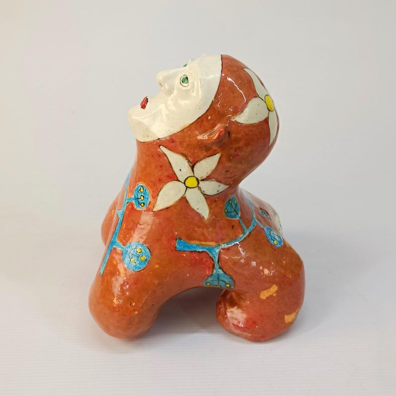 May include: A ceramic sculpture of a stylized human figure with a red, white, and blue floral design. The figure is sitting with its head tilted back and its eyes closed.