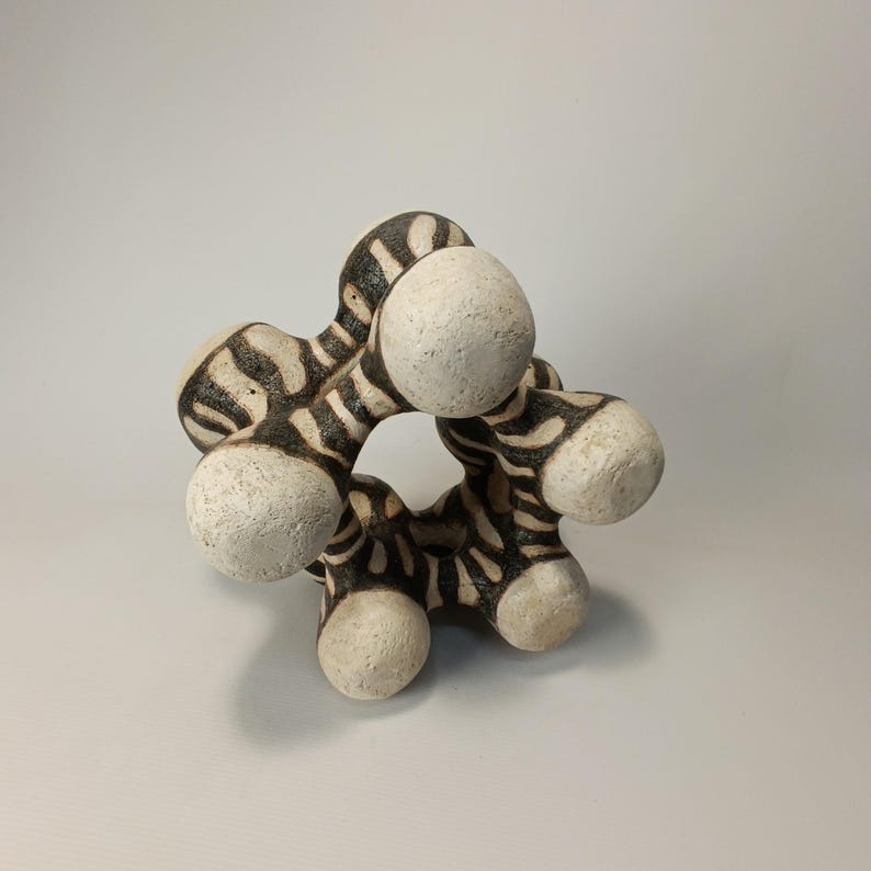 May include: A ceramic sculpture with a unique, abstract design. The piece features a circular form with six rounded, off-white spheres connected by striped, dark brown and cream-colored segments. The sculpture has a textured surface and is displayed against a plain background.