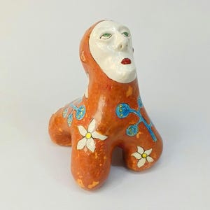 May include: A ceramic sculpture of a creature with a human-like face and a body covered in orange, white, and blue floral designs.