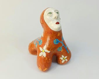 May include: A ceramic sculpture of a creature with a human-like face and a body covered in orange, white, and blue floral designs.