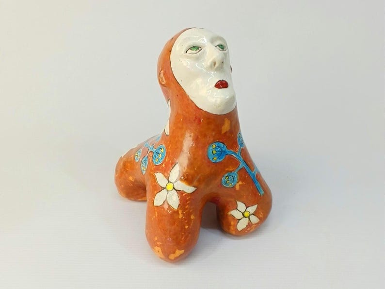 May include: A ceramic sculpture of a creature with a human-like face and a body covered in orange, white, and blue floral designs.