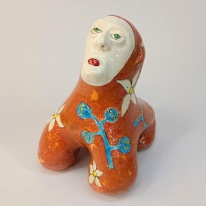 May include: A ceramic sculpture of a stylized human figure with a white face, red lips, and green eyes. The figure is wearing a red and orange outfit with white flowers and blue branches.
