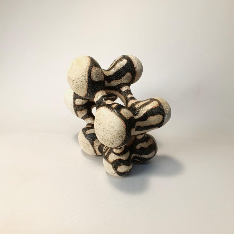 May include: Abstract ceramic sculpture with a complex, interconnected form. The piece features a cream-colored base with bold black zebra-like stripes. The rounded shapes create a visually interesting and modern art piece.