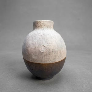 Może przedstawiać: Sferyczny wazon ceramiczny o dwukolorowym wzorze, z teksturowaną górną częścią w kolorze złamanej bieli oraz ciemnobrązową, szorstką podstawą. Na górnej białej części wazonu widoczna jest mała okrągła pieczątka ze stylizowanym symbolem.