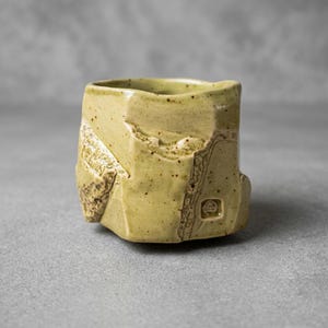 May include: A hand-thrown ceramic cup with a pale yellow, speckled glaze. The vessel features an irregular, faceted exterior surface with deep, textured indentations and a small, square-shaped stamped mark near the base of the cup.