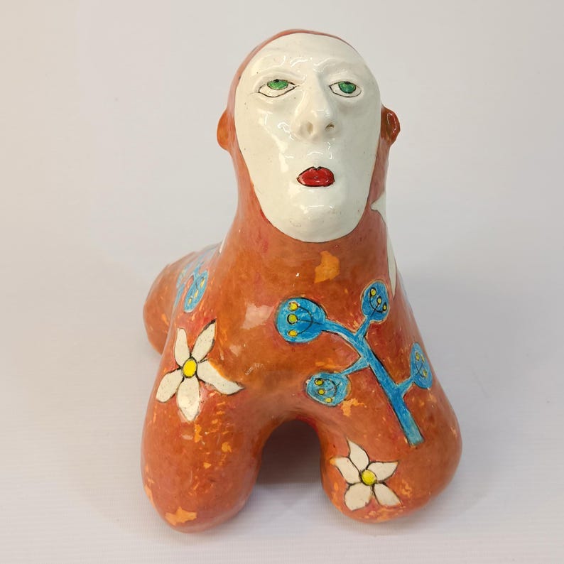 May include: A ceramic sculpture of a stylized human figure with a white face, green eyes, and red lips. The figure is painted in orange, white, blue, and yellow with floral designs.