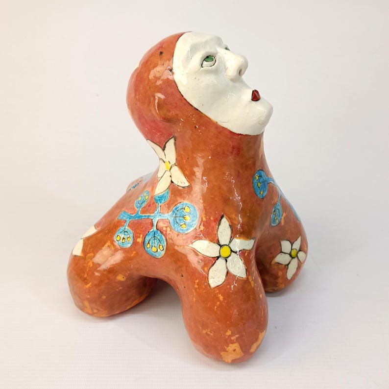 May include: A ceramic sculpture of a person with a white face and red lips, wearing a red and orange outfit with white flowers and blue leaves. The sculpture is standing on three legs.